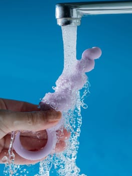 Woman holding lilac anal beads under running water on blue background. Sex toy hygiene concept