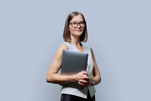 Young confident woman with laptop computer in hands on gray studio background. Smiling female with glasses looking at camera. Using mobile application for work business study leisure technology people