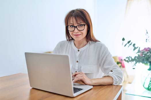 Middle aged business woman working at desk in home office, looking at camera with laptop computer. Work, remote business, job, online teaching, financial legal control, mature people lifestyle concept