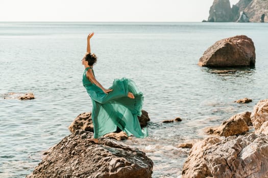 Woman green dress sea. Female dancer in a long mint dress posing on a beach with rocks on sunny day. Girl on the nature on blue sky background