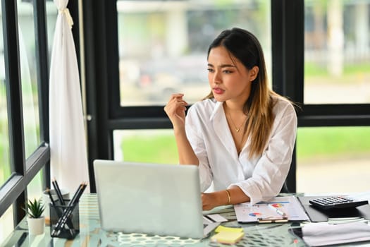 Thoughtful female economist sitting at desk and looking away, thinking of analysis planning business vision.