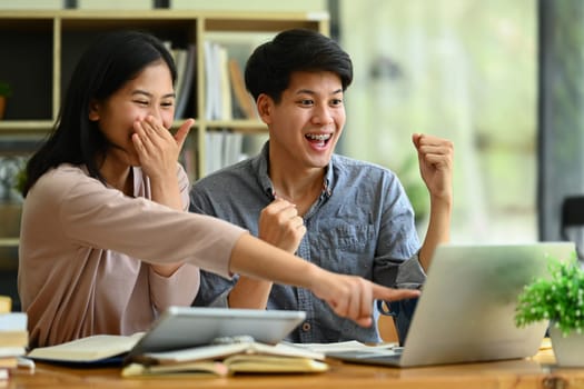 Overjoyed college students looking at laptop screen celebrating success, exam results together.