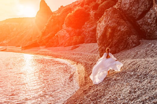 Mysterious woman silhouette long hair walks on the beach ocean water, sea nymph wind listens to the wave. Throws up a long white dress, a divine sunset. Artistic photo from the back without a face.