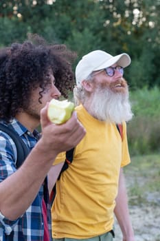 two friends of different ages go hiking with backpacks, and an Arab guy with black hair eats a green apple with appetiteagainst the background of a forest, High quality photo