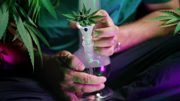 Man smoking marijuana from water bong in neon light at home. Cannabis, hemp. High quality photo
