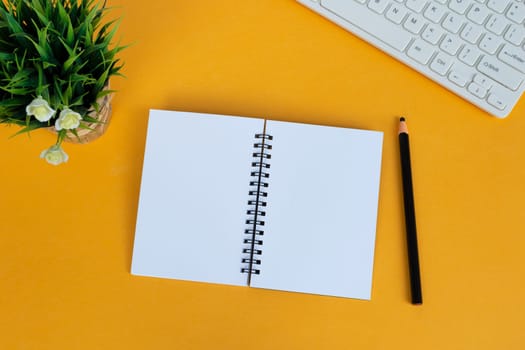 Directly above view of office desk with white computer keyboard, pen, notepad, and potted plant on yellow background. Copy space. Flat lay.
