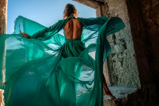 Rear view of a happy blonde woman in a long mint dress posing against the backdrop of the sea in an old building with columns. Girl in nature against the blue sky