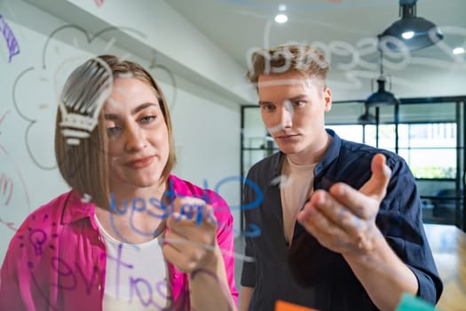 Couple of businesspeople discussing and brainstorming about solving marketing problem in front of glass board at business meeting by using mind map and sticky notes. Working together. Immaculate.