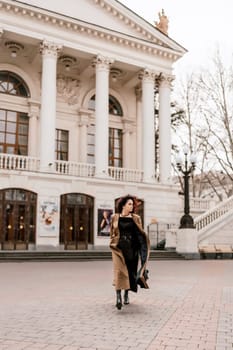 Woman street lifestyle. Image of stylish woman walking through European city on sunny day. Pretty woman with dark flowing hair, dressed in a beige raincoat and black, walks along the building