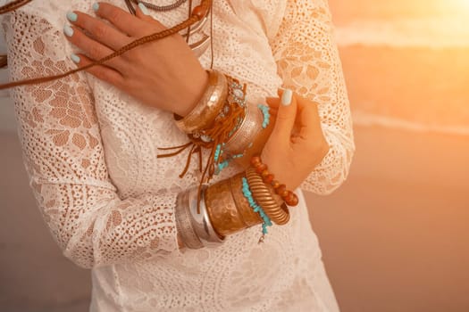 Model in boho style in a white long dress and silver jewelry on the beach. Her hair is braided, and there are many bracelets on her arms