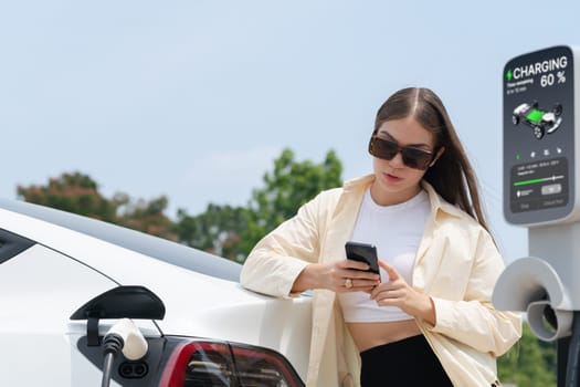 Young woman use smartphone to pay for electricity at public EV car charging station green city park. Modern environmental and sustainable urban lifestyle with EV vehicle. Expedient