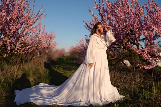 Woman blooming peach orchard. Against the backdrop of a picturesque peach orchard, a woman in a long white dress enjoys a peaceful walk in the park, surrounded by the beauty of nature