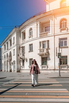 Woman city road crossing. Stylish woman in a hat crosses the road at a pedestrian crossing in the city. Dressed in white trousers and a jacket with a bag in her hands