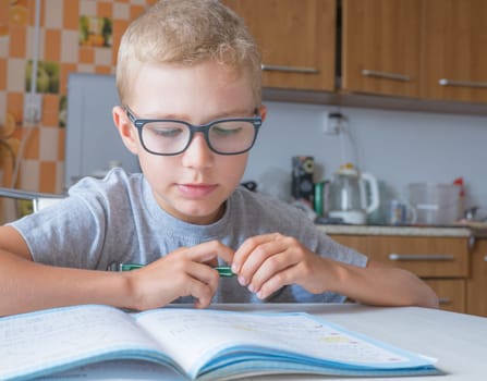 Tired sad little child boy sits at a table in the kitchen and writes homework. Boring lessons.