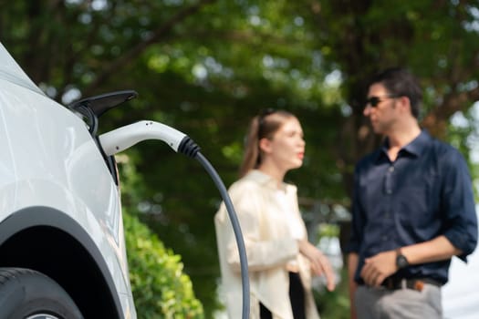 Young couple recharge electric car's battery from charging station in outdoor green city park in springtime. Rechargeable EV car for sustainable environmental friendly urban travel lifestyle.Expedient