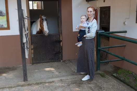 Mom holds her baby - stand next to a stall with a horse. Mid shot
