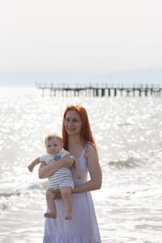 Happy mother on vacation - smiling woman standing on the beach holding her little baby son. Portrait
