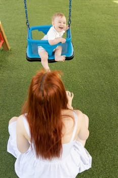 A little funny boy on swings on an outside playground with his ginger mother. Mid shot