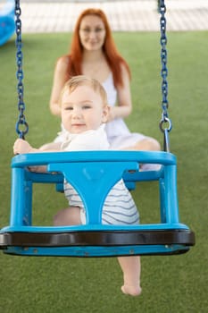 A little boy on swings on an outside playground with his ginger mother - looking in the camera. Mid shot