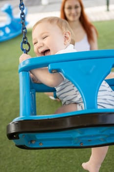 A little laughing boy on swings on an outside playground with his ginger mother - looking in the camera. Mid shot