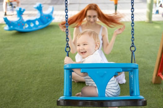 A little laughing boy on swings on an outside playground with his ginger mother. Mid shot