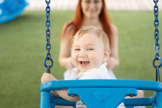A little laughing baby on swings on an outside playground with his ginger mother. Mid shot