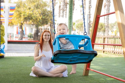 Funny baby swings on swings in playground and his mom sits near him. Mid shot
