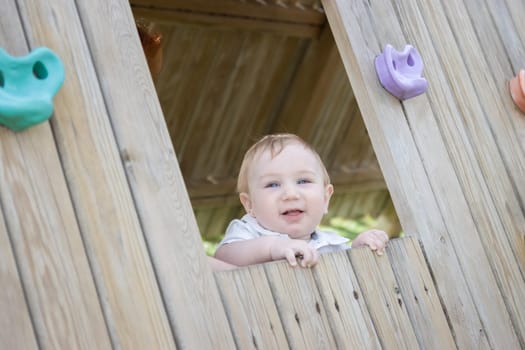 Funny baby sits in a wooden house on the playground. Mid shot