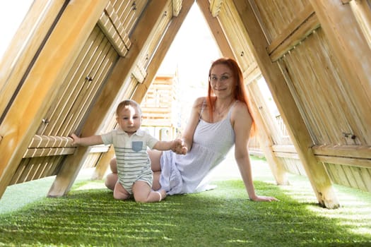 Cute baby and his mother sits under a wooden roof on the playground - looking in the camera. Mid shot