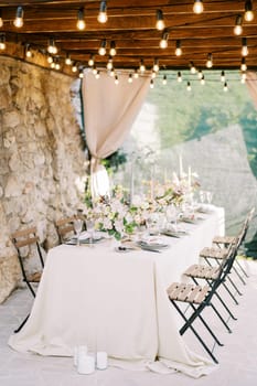 Laid festive table with chairs stands against a stone wall under a wooden canopy with glowing garlands. High quality photo