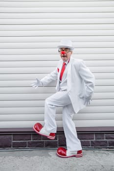 An elderly man in a white suit, huge boots and a clown nose walks funny