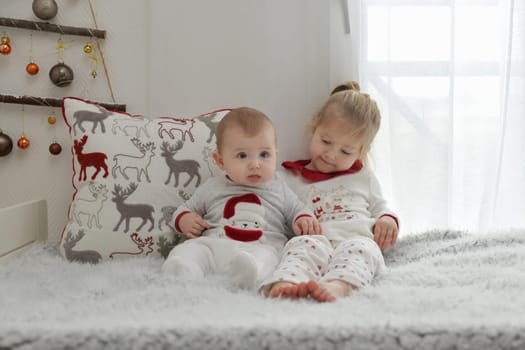 Brother and sister in Christmas pajamas are sitting on the bed with decorations.