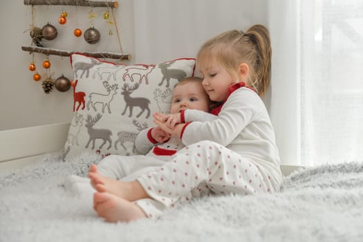 Brother and sister in Christmas pajamas are sitting on the bed with decorations.