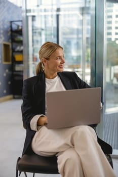 Pretty female entrepreneur working on laptop sitting in modern office hall and looks away