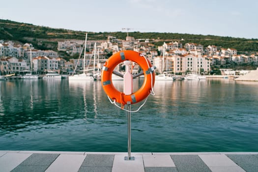Orange lifebuoy on a stand on a pier against the backdrop of a colorful resort town with moored yachts. High quality photo