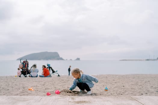 Little girl digs a hole in the sand while squatting against the background of mothers with children sitting on the shore. High quality photo