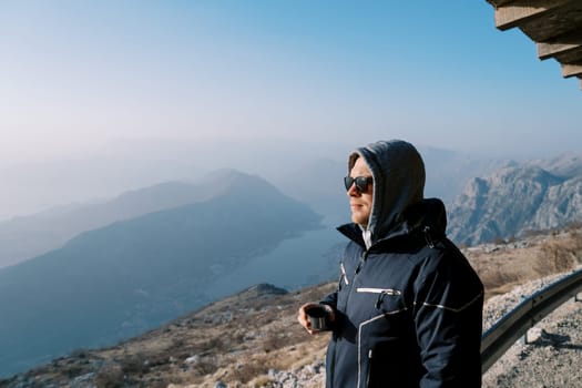 Man in a hooded jacket with a thermos mug stands in the mountains above the bay. Side view. High quality photo