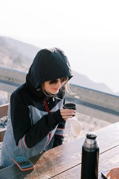 Girl in a jacket with a hood sits at a table in the mountains and drinks coffee with a mug from a thermos. High quality photo