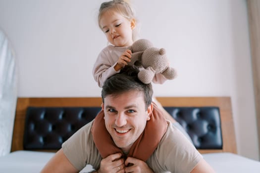 Little girl with a teddy bear sits on the shoulders of her father, who lies on his stomach on the bed. High quality photo