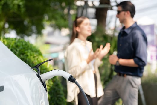 Young couple recharge electric car's battery from charging station in outdoor green city park in springtime. Rechargeable EV car for sustainable environmental friendly urban travel lifestyle.Expedient