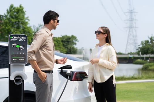 Young couple recharge EV car battery at charging station connected to power grid tower electrical industrial facility as electrical industry for eco friendly vehicle utilization. Expedient