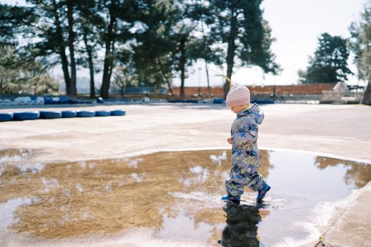 Little girl in rubber boots and overalls walks through a large puddle on the pavement. Side view. High quality photo