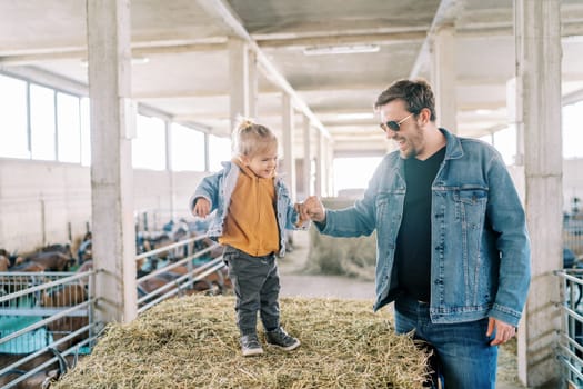 Smiling dad holding the hand of a little girl standing on a bale of hay at the farm. High quality photo