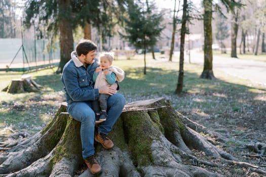 Dad with a little girl on his knees sits on a huge stump in a sunny spring park. High quality photo