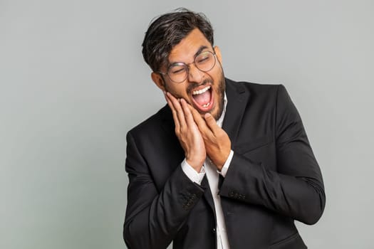 Dental problems. Indian businessman touching cheek, closing eyes with expression of terrible suffer from painful toothache, sensitive teeth, cavities. Male Arabian man guy isolated on gray background