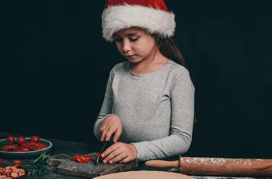 Little girl in santa hat cuts with a big black knife cherry tomatoes on a gray wooden board, next to pizza dough, rolling pin and Christmas decor on a dark background, close-up side view. Cooking concept.