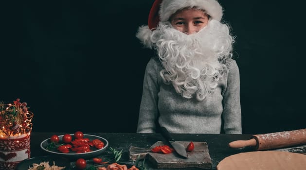 Caucasian teenage girl in a Christmas hat with a beard sits cheerful at the table with cherry tomatoes on a gray wooden board, next to pizza dough, rolling pin and Christmas decor on a dark background, close-up side view. Cooking concept.