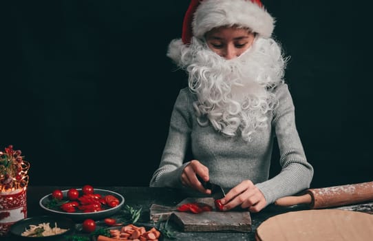 Caucasian teenage girl in a santa hat with a beard cuts with a big black knife cherry tomatoes on a gray wooden board, next to pizza dough, rolling pin and Christmas decor on a dark background, close-up side view. Cooking concept.
