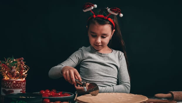 A Caucasian little girl cuts a Christmas tree with a round moon knife on a pizza dough, and next to a rolling pin, sliced cherry tomatoes, ketchup and a decoration in a mug with a sweater on a dark background, close-up side view.
