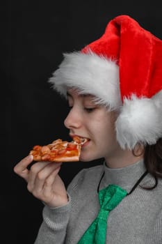 Happy caucasian teenage girl wearing a santa claus hat holds a slice of pizza in her hand and bites with her mouth with copy space for your text on a black background, close-up side view.